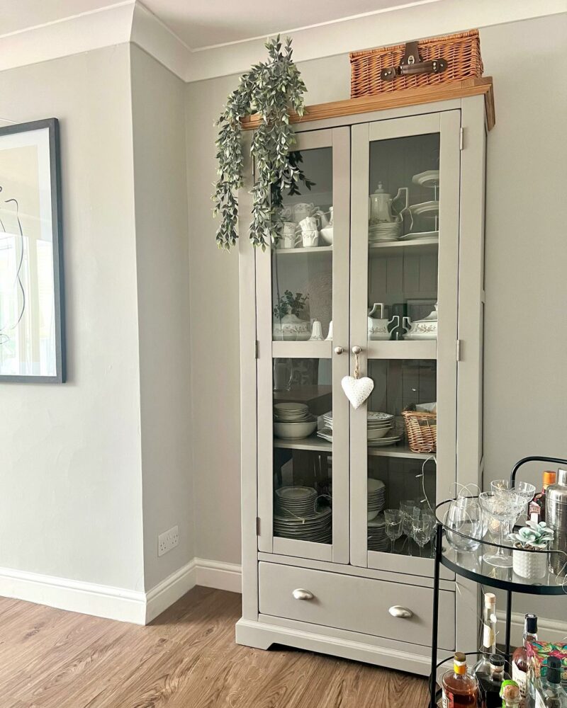 Grey painted glass-fronted display cabinet in an alcove with a barcart next to it.