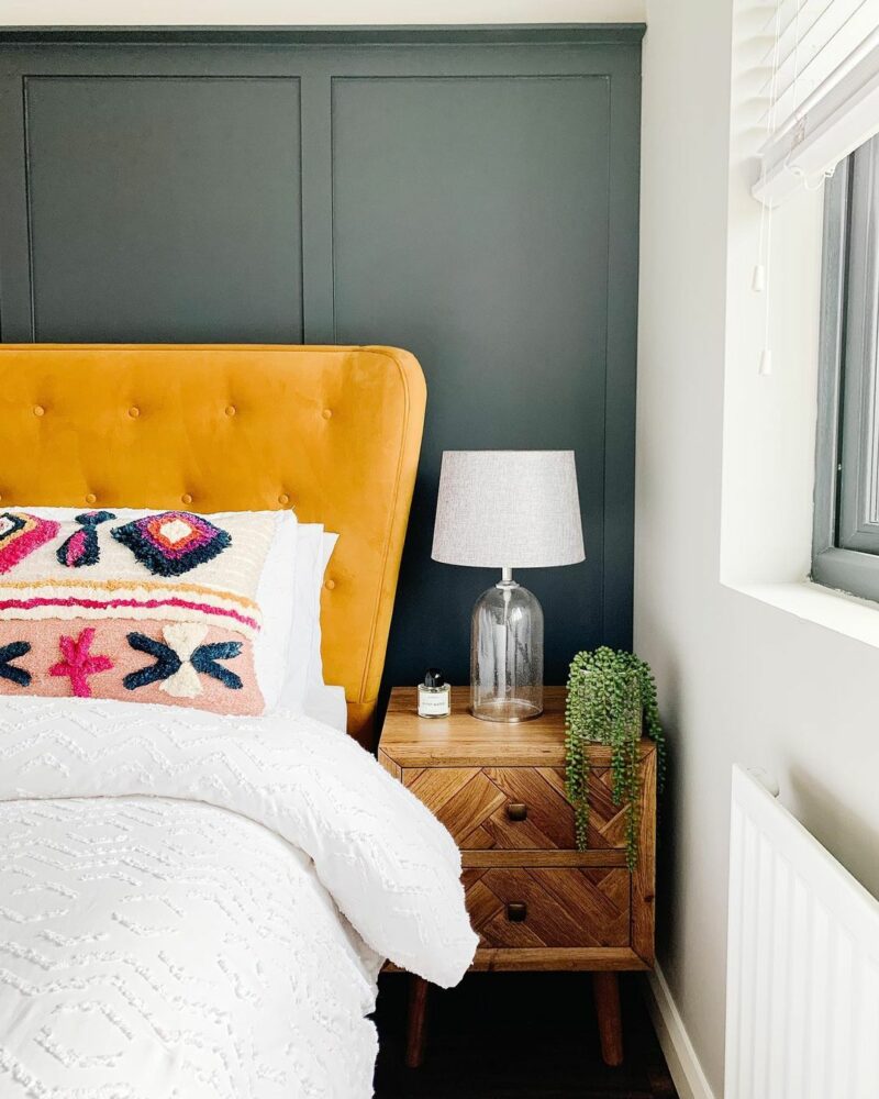 Bedroom with a dark green/grey wall featuring a contrasting yellow velvet bed, colourful cushions and a Parquet solid oak bedside table.