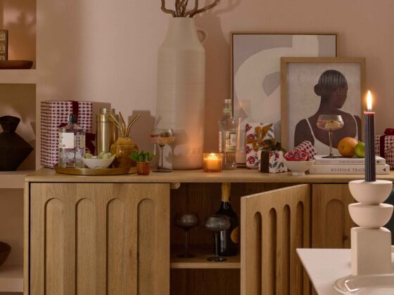A sideboard-dining room furniture-arch detail wooden sideboard-abstract artwork-pink blush walls-cocktail shaker-wrapped presents-metallic tray-spirits-mixers-straws-cocktail glasses-fresh mint and cucumber-oversized cream vase-sculptural candle holder-lit candles-built in shelving