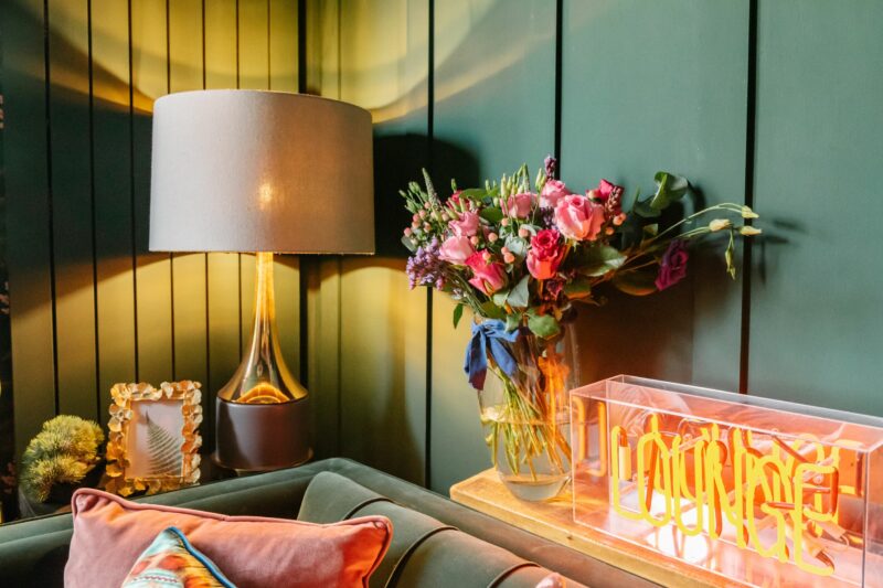 Cosy dark green living room with Oak Furnitureland Waldorf lamp, vase of flowers and neon light.