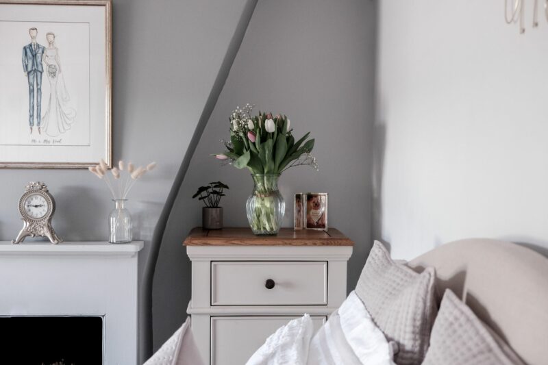 Oak Furnitureland Shay grey painted tallboy in an elegant bedroom with a vase of flowers & photos.