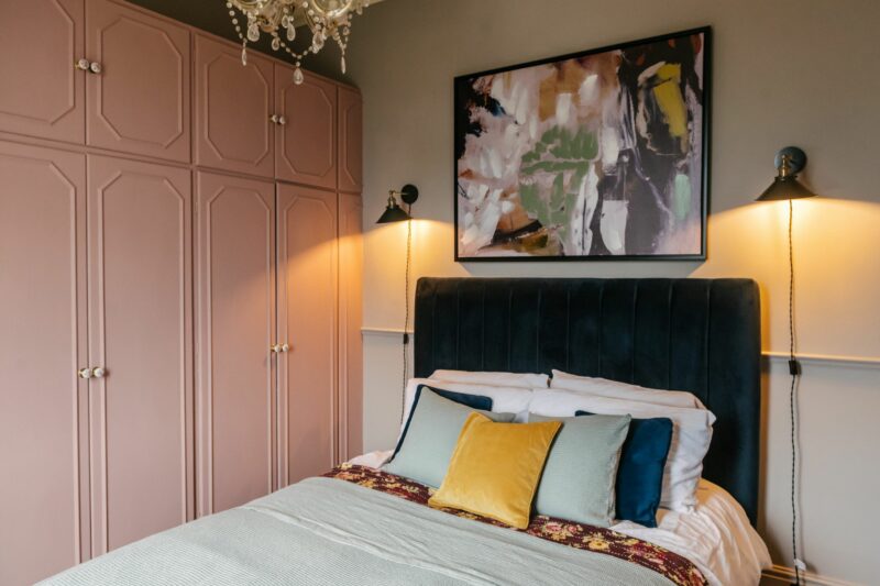 Velvet upholstered bed covered in cushions in a bedroom with pale pink fitted wardrobes and tonal artwork displayed above the bed.