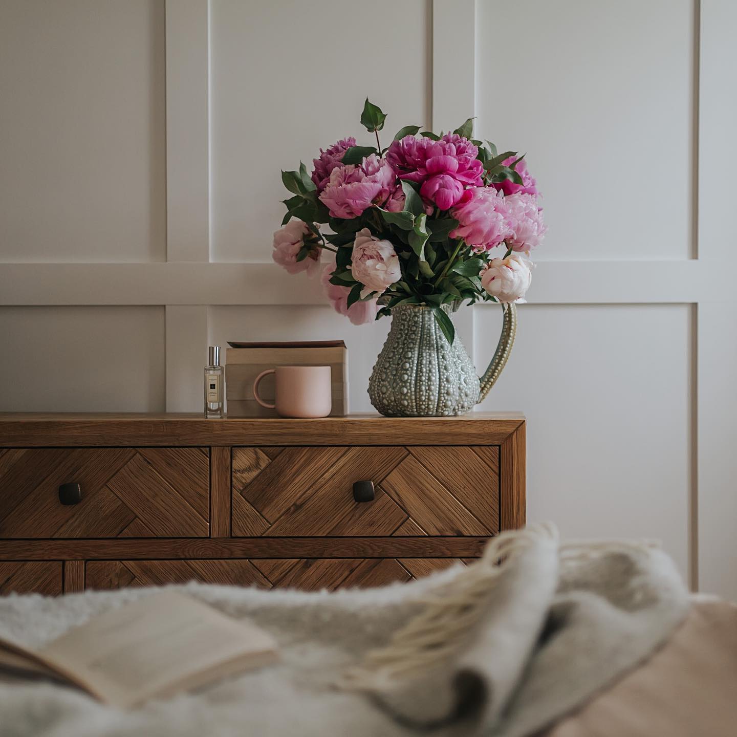Parquet chest of drawers in a cream bedroom with a jug of pink flowers on top.