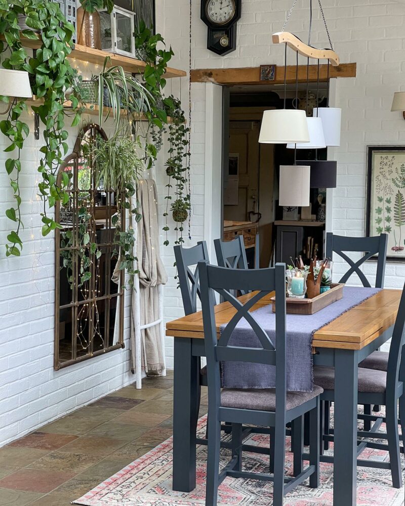 A painted dark blue and hardwood Oak Furnitureland Highgate dining set with a light blue fabric runner sits in a naturally lit room. Houseplants sit on a wooden wall shelf that runs parallel to the dining set and hang down in front of a large window pane mirror.