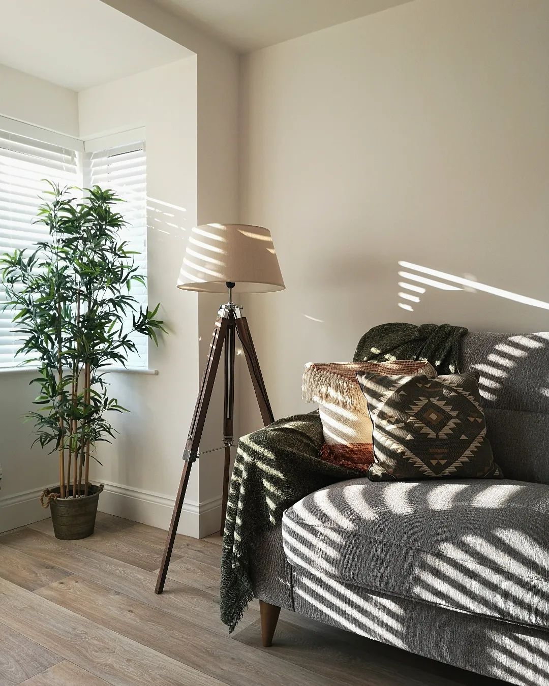 Sunlight spills through slatted blinds into a living room. A large houseplant and floor lamp stand next to a grey Oak Furnitureland Jensen sofa that has been styled with a cosy throw and scatter cushions.