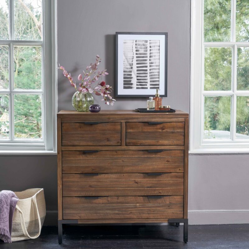 A chest of drawers-bedroom furniture-mauve walls-dark wooden drawers-metal handles and feet-dark grey floor-cotton tote bag-purple scarf-monochrome wall art-vase of leaves-tray of perfume