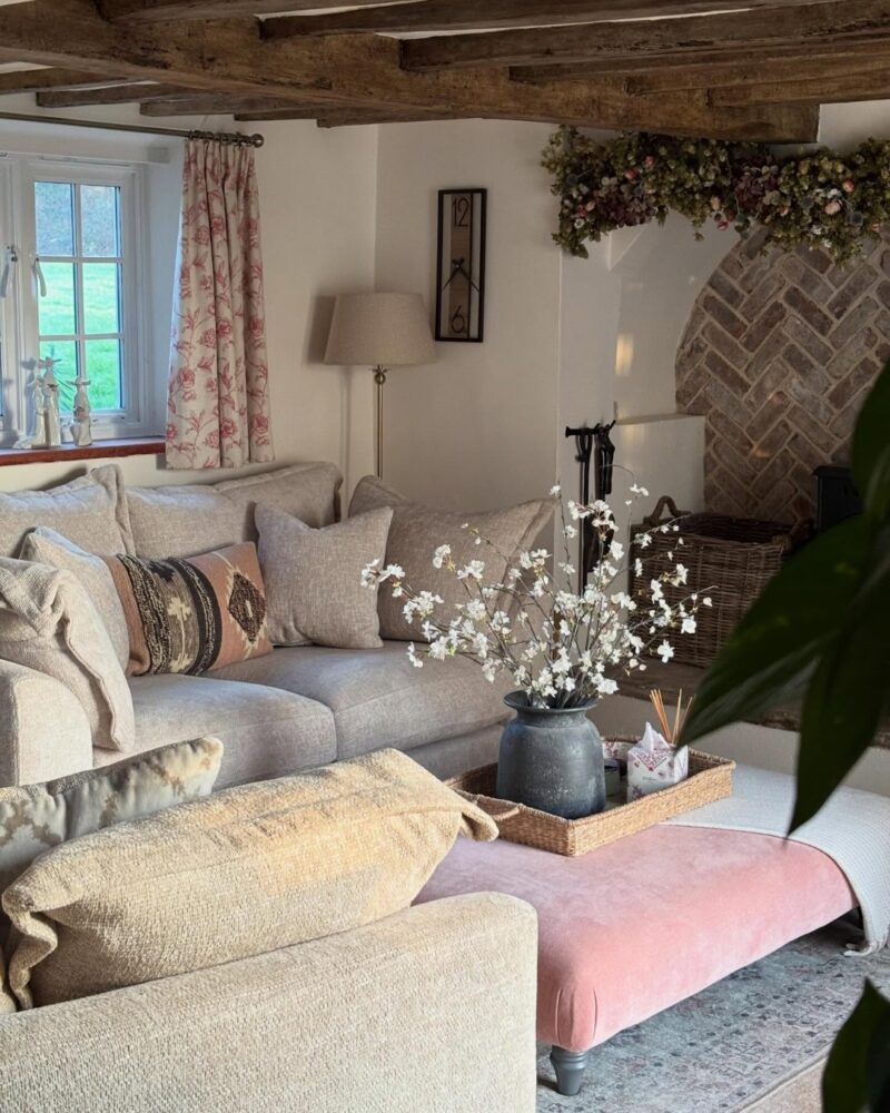 A couple of sofas and a footstool-living room furniture-beige sofas-patterned pink cushions-neutral cushions-soft pink footstool-woven tray-charcoal vase with dried flowers-vintage style rug-floral curtains-ceiling with wooden beams-beige walls-woven wood basket-neutral floor standing lamp