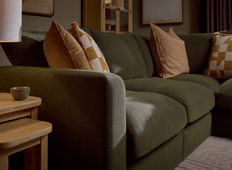 A sofa and nest of tables-living room furniture-dark green corner modular sofa-caramel coloured cushions-geometric cushions-wooden nest of tables-textured neutral rug-wood and glass display cabinet-cream table lamp