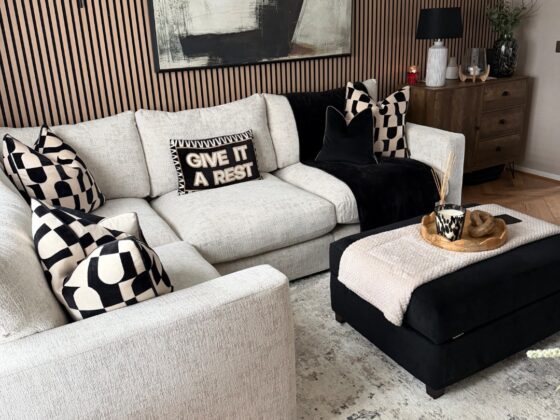A sofa, footstool, and sideboard-living room furniture-beige corner modular sofa-black footstool-black and white geometric cushions-black throw-neutral abstract rug-wooden floor-wooden panelling on feature wall-large abstract monochrome painting-black wall lights-beige wall-dark wood sideboard-wooden scallop tray-cream throw