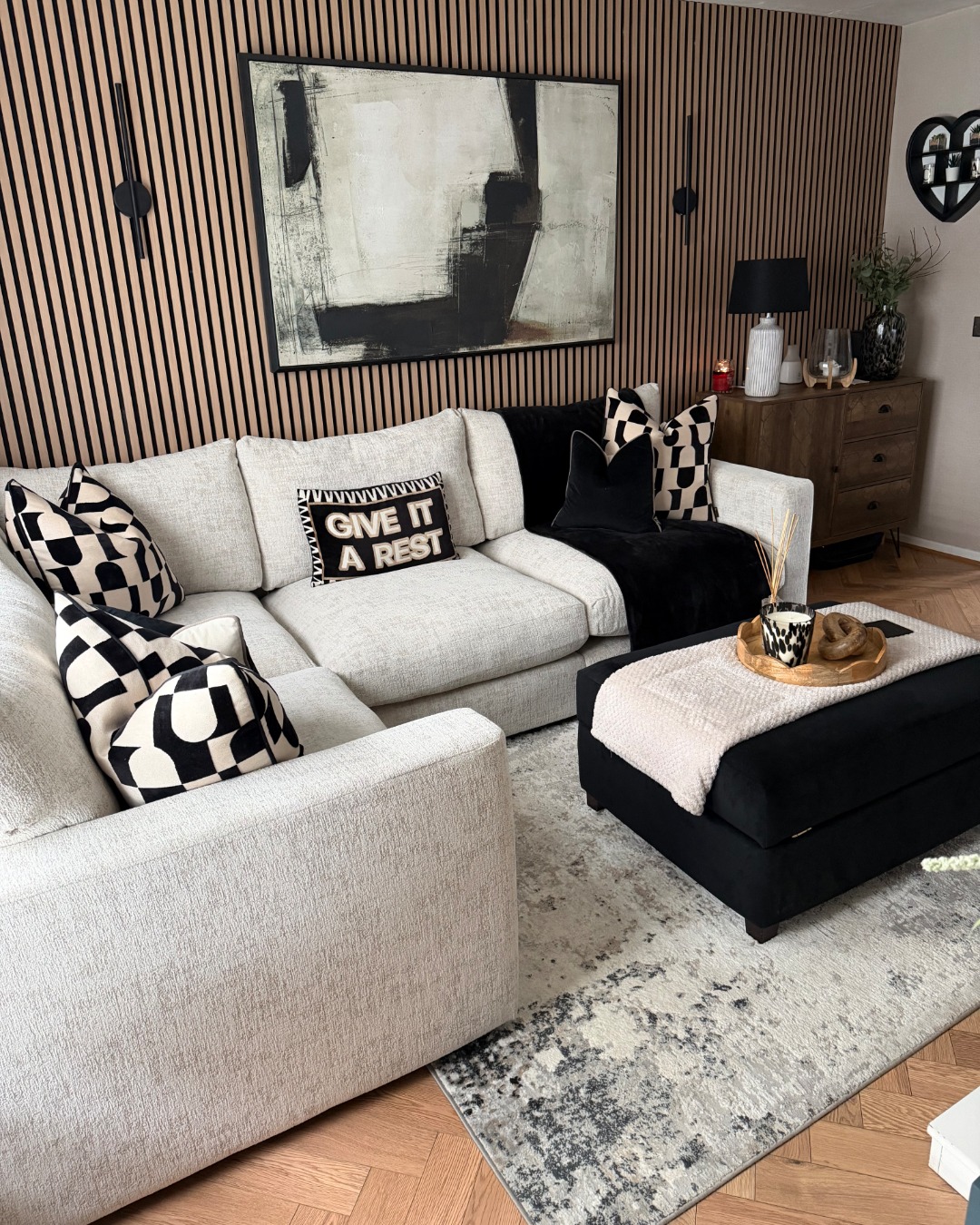 A sofa, footstool, and sideboard-living room furniture-beige corner modular sofa-black footstool-black and white geometric cushions-black throw-neutral abstract rug-wooden floor-wooden panelling on feature wall-large abstract monochrome painting-black wall lights-beige wall-dark wood sideboard-wooden scallop tray-cream throw
