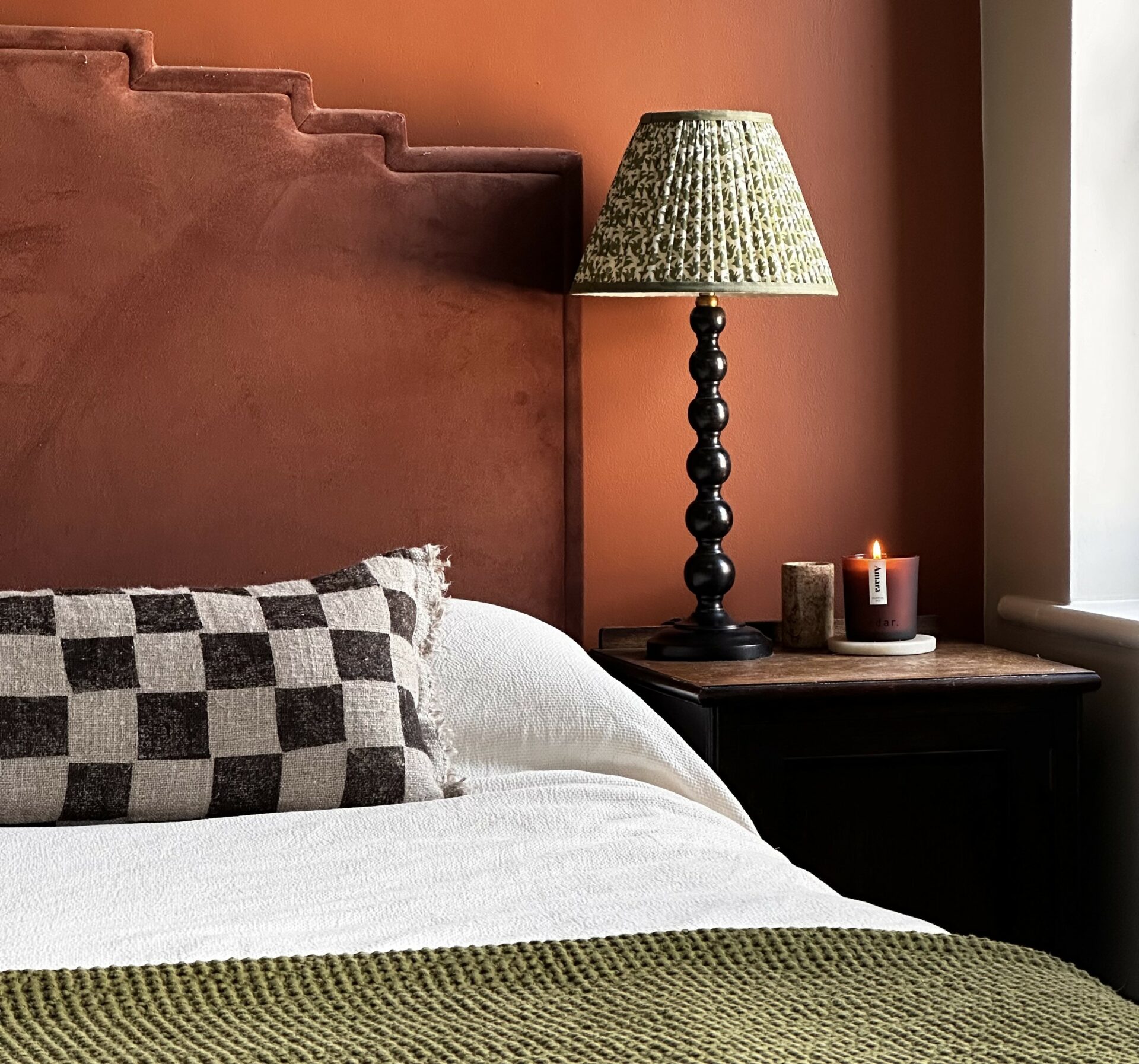 A bed, bedside tables, table lamps-bedroom furniture-terracotta walls-amber glass candle-checkered cushion-bobbin lamps-dark wooden bedside table-rust red velvet headboard- green throw-white bed linen