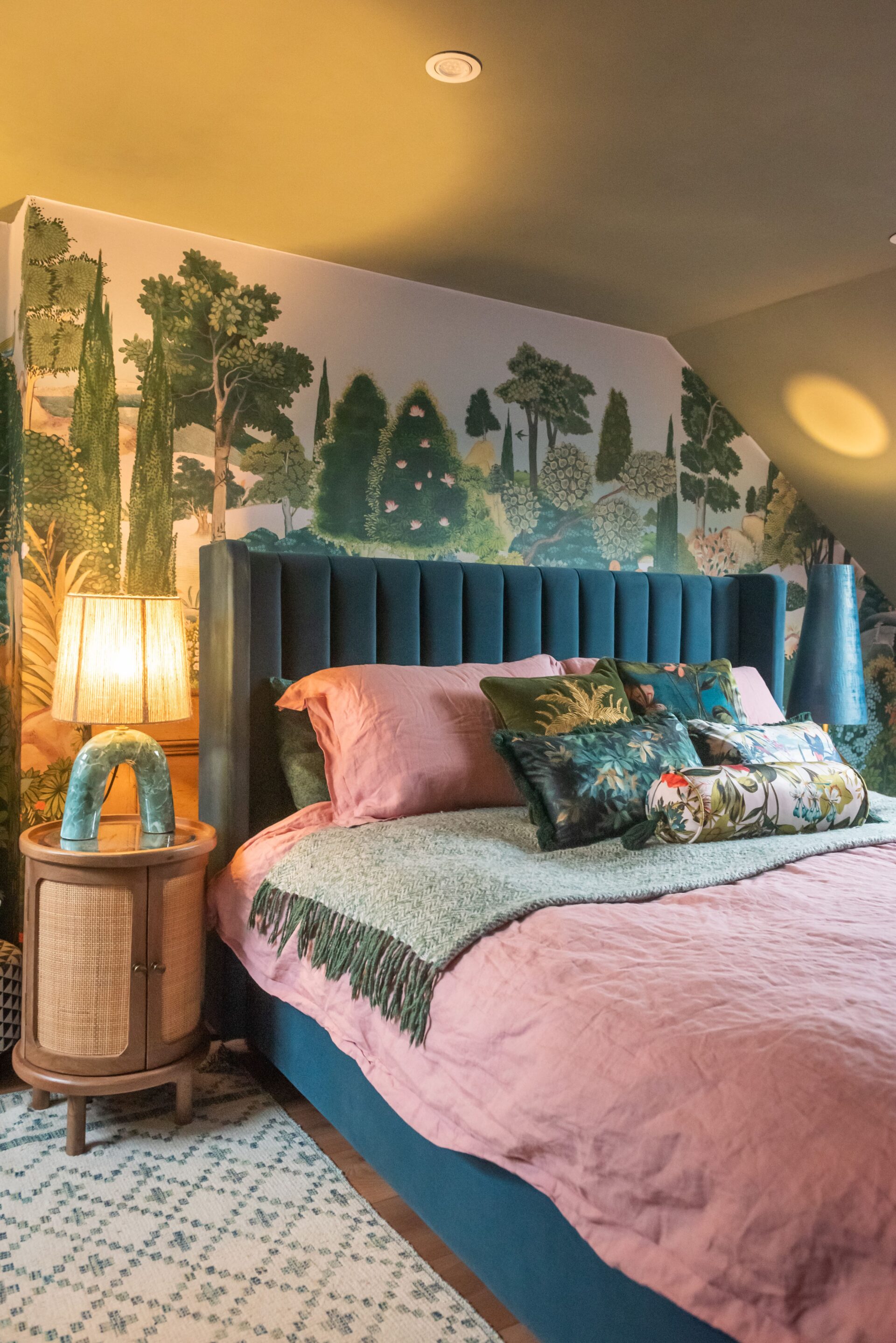 A bed and bedside table-bedroom furniture-forest wall mural-teal upholstered bed-pink bedding-statement lamp-wooden bedside table-storage bed