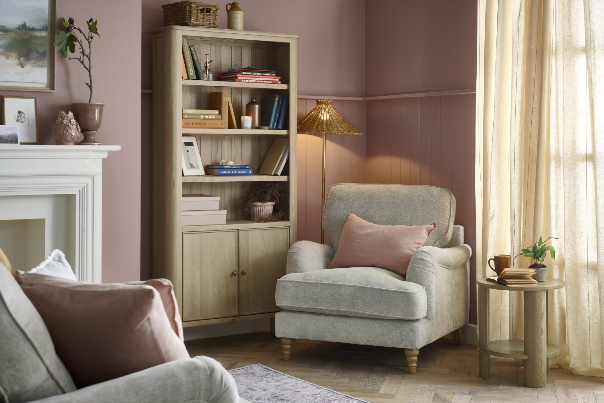 An armchair, sofa, and bookcase-living room furniture-dusky pink walls-parquet wooden floor-vintage pattern rug-grey sofa-grey armchair-pink linen cushion-fireplace-potted plants-landscape wall art-round oak side table-smoked oak bookcase-colourful books-cream curtains