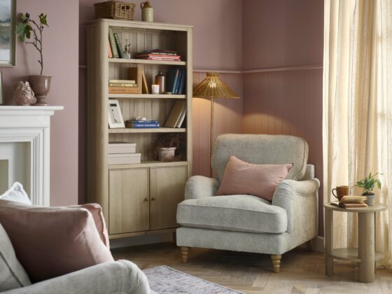 An armchair, sofa, and bookcase-living room furniture-dusky pink walls-parquet wooden floor-vintage pattern rug-grey sofa-grey armchair-pink linen cushion-fireplace-potted plants-landscape wall art-round oak side table-smoked oak bookcase-colourful books-cream curtains