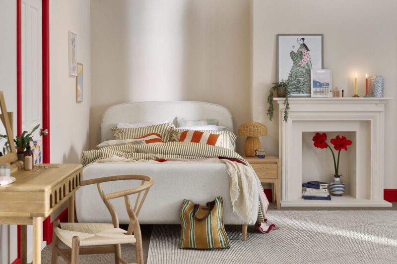 A bed, bedside table, and fireplace-bedroom furniture-green striped bedding-red striped cushion-blue striped cushion-red and pink throw-neutral upholstered bed-pale wooden bedside table-wicker mushroom lamp-neutral walls-colourful candles-abstract wall art-pale wooden dressing table with arched drawers-wishbone style chair