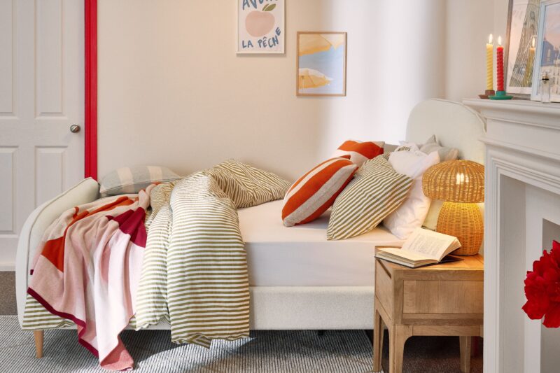 A bed and bedside table-bedroom furniture-green striped bedding-red striped cushion-blue striped cushion-red and pink throw-neutral upholstered bed-pale wooden bedside table-wicker mushroom lamp-neutral walls-colourful candles-abstract wall art
