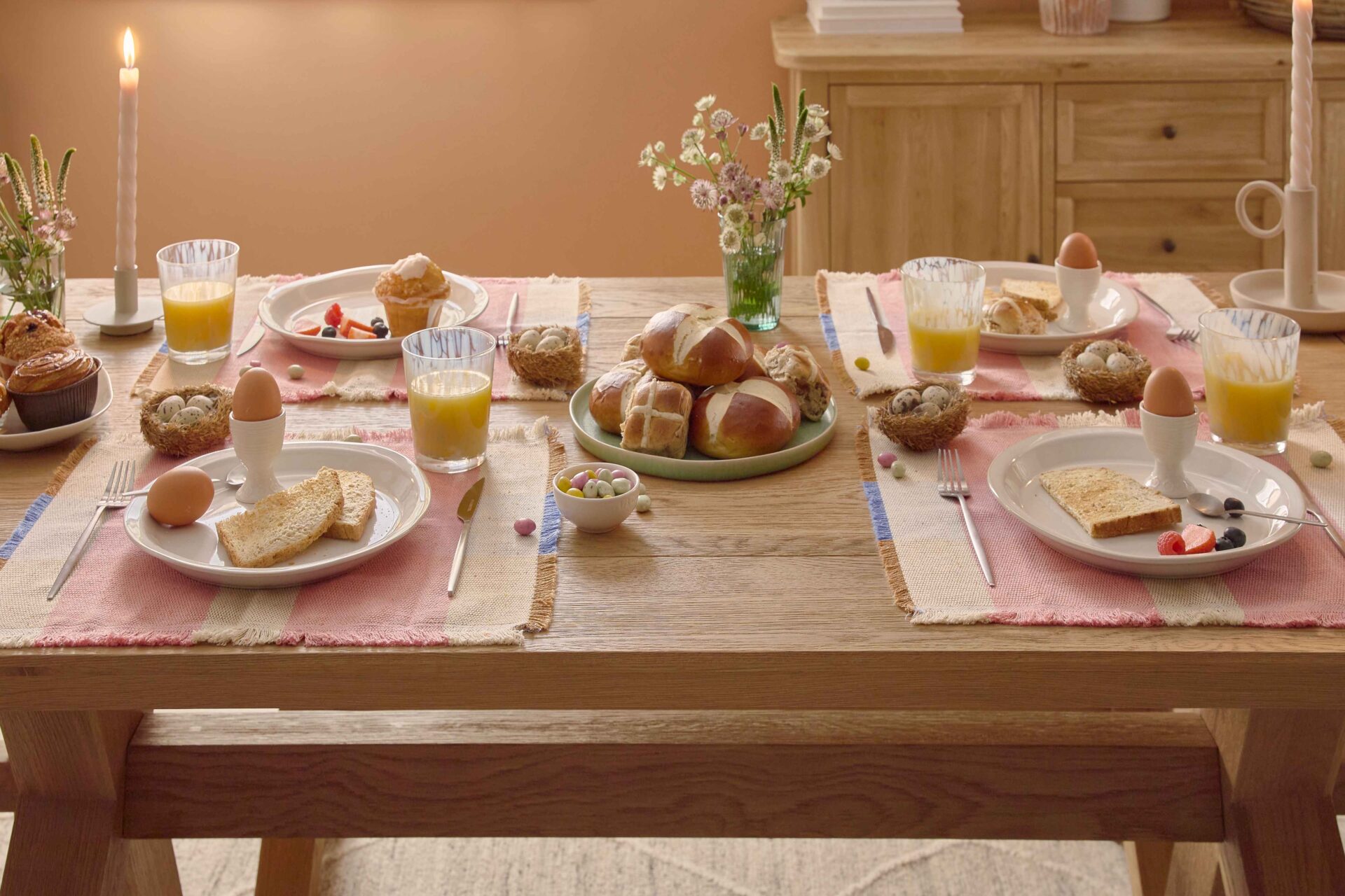 A dining table set-dining room furniture-natural oak dining table and benches-soft terracotta walls-neutral rug-wooden sideboard-Easter tablescape-colourful placemats-wild flowers in vases-hot cross buns-fresh orange juice in glasses-boiled eggs-fresh fruit-bowls of mini eggs