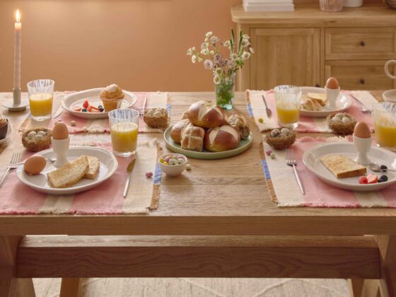 A dining table set-dining room furniture-natural oak dining table and benches-soft terracotta walls-neutral rug-wooden sideboard-Easter tablescape-colourful placemats-wild flowers in vases-hot cross buns-fresh orange juice in glasses-boiled eggs-fresh fruit-bowls of mini eggs