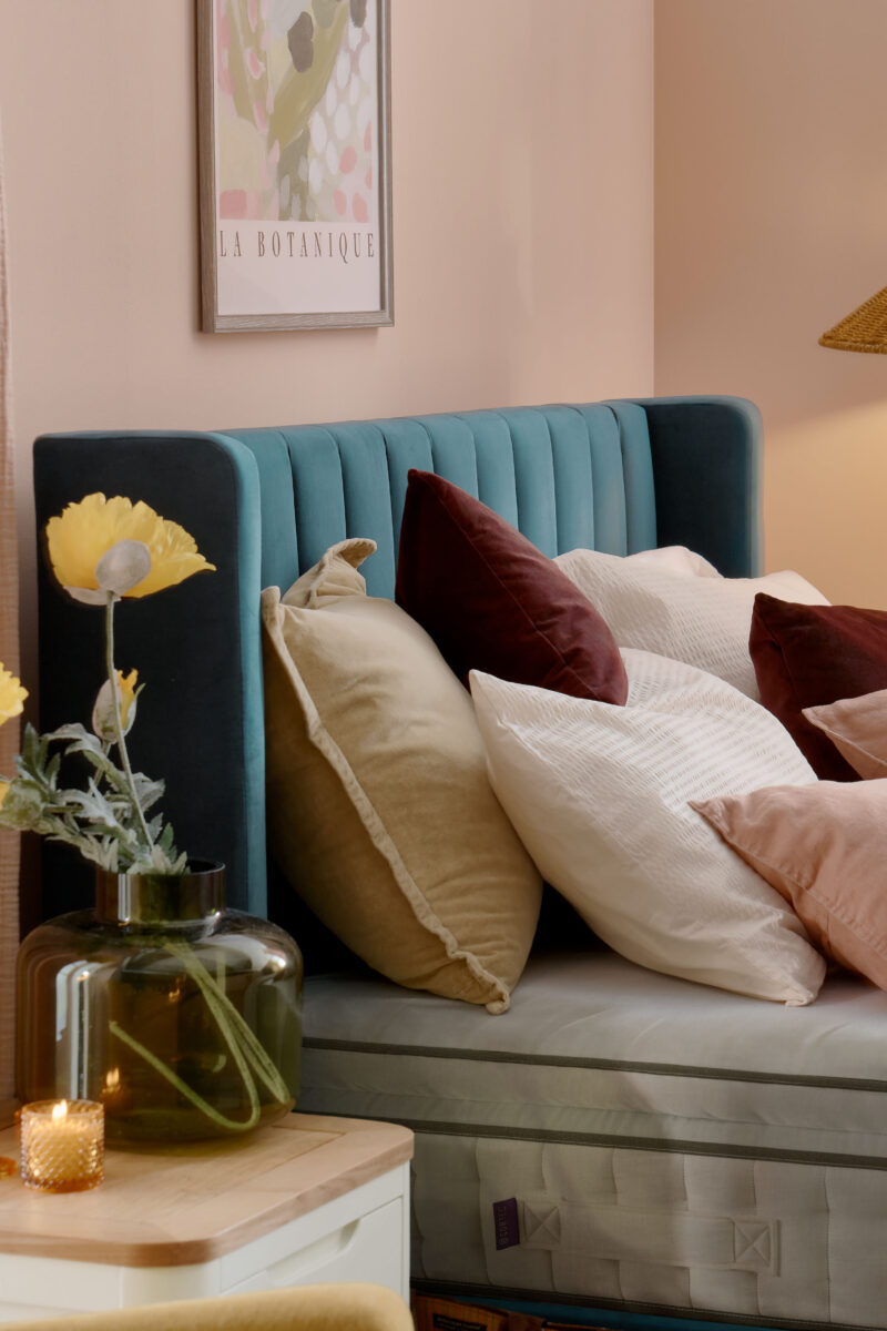 A bed and bedside table-bedroom furniture-teal upholstered bed-layered cushions-plain mattress-pink walls-green vase with flowers-candle light-cream and wood bedside table-floral wall art
