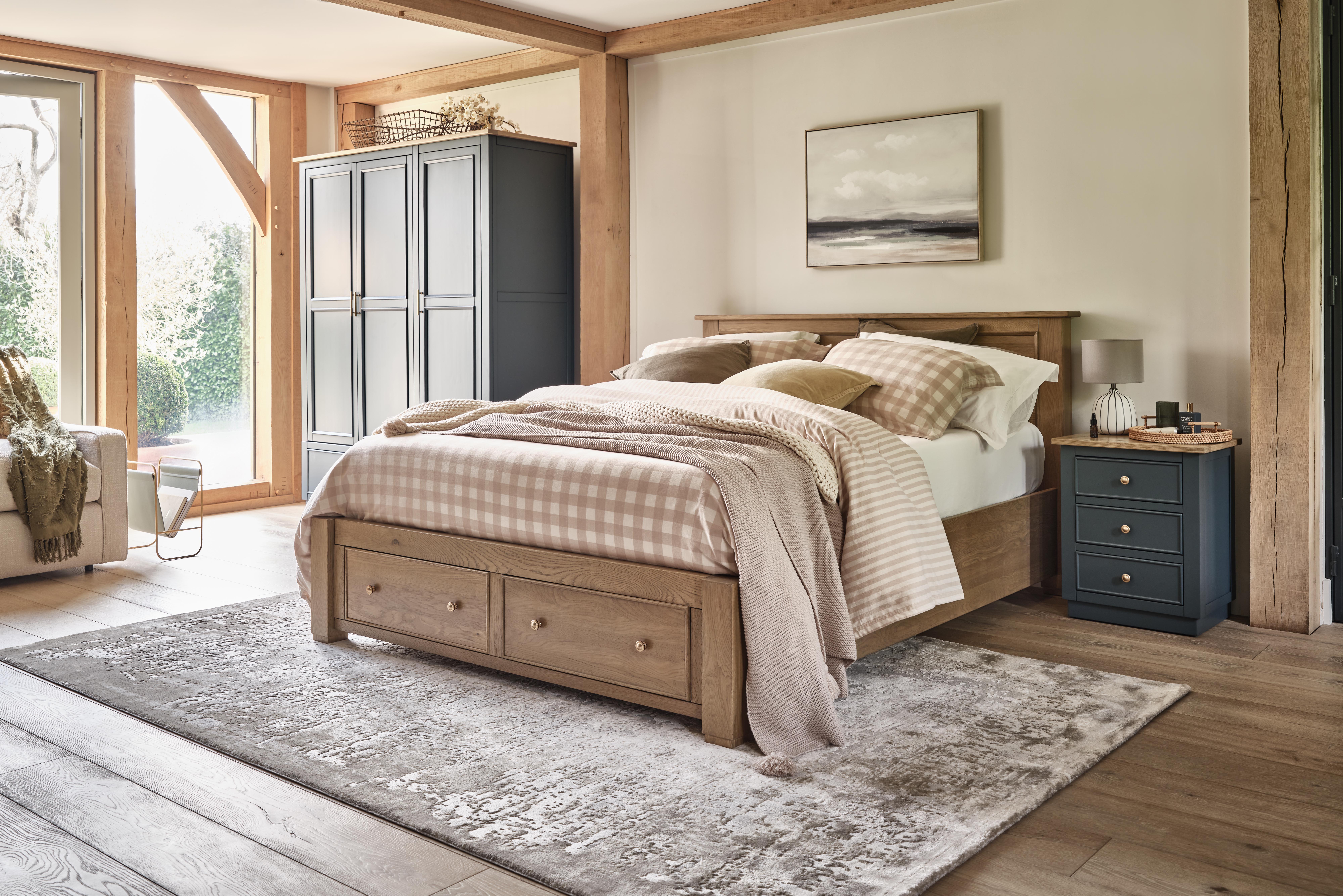 A bed, wardrobe, and bedside table-bedroom furniture-wooden bed with storage drawers-neutral bedding-check bed linen-navy blue cabinetry-wooden floor-textured neutral rug-neutral walls-exposed beams-blue bedside table with drawers-blue triple wardrobe