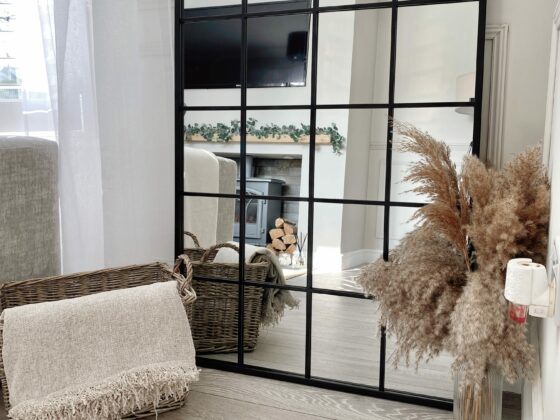 A mirror-living room furniture-black windowpane floor mirror-dried grass in glass vase-wicker storage basket-cream throw-neutral walls-log burner-white washed wooden floorboards