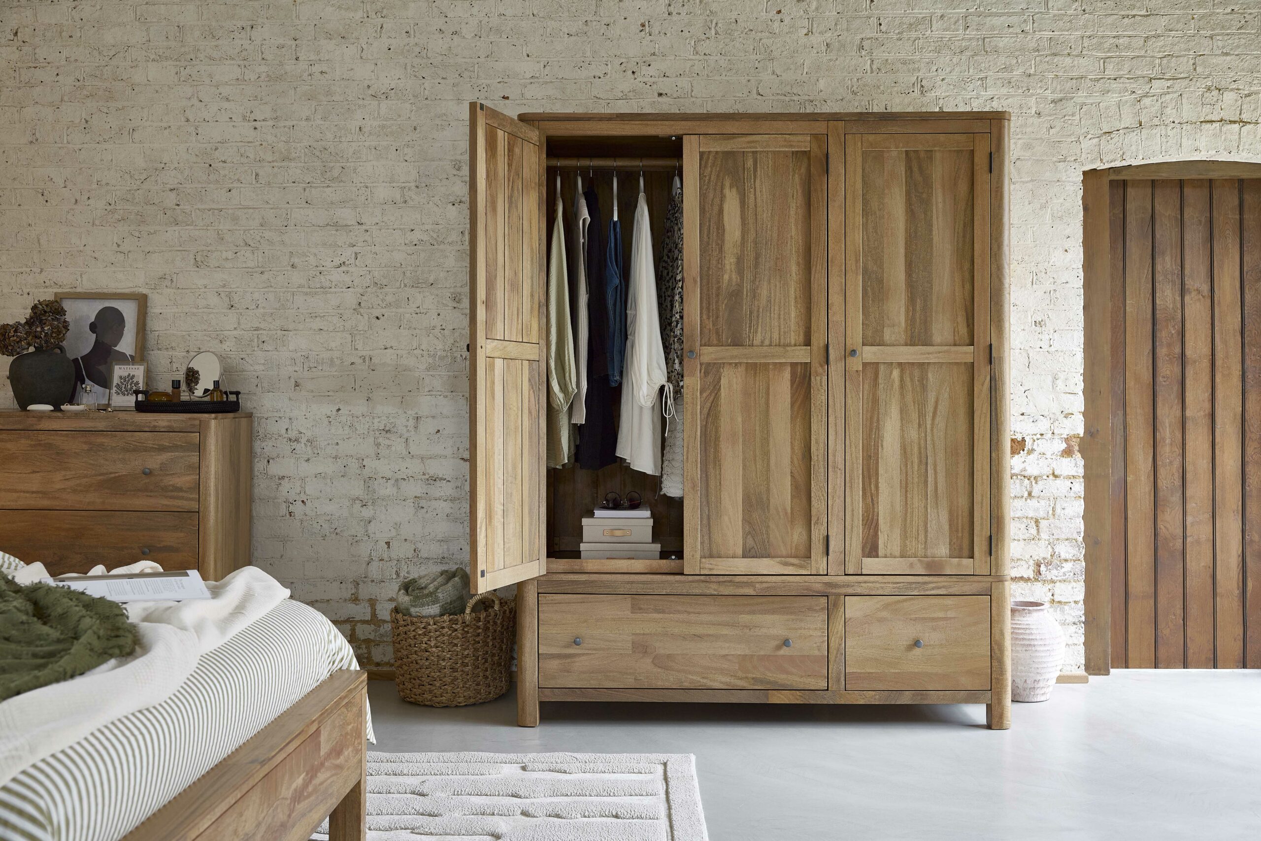 A wardrobe, bed, and chest of drawers-bedroom furniture-mango wood wardrobe-textured neutral rug-neutral painted brick wall-wooden door-polished concrete floor-green striped bedding