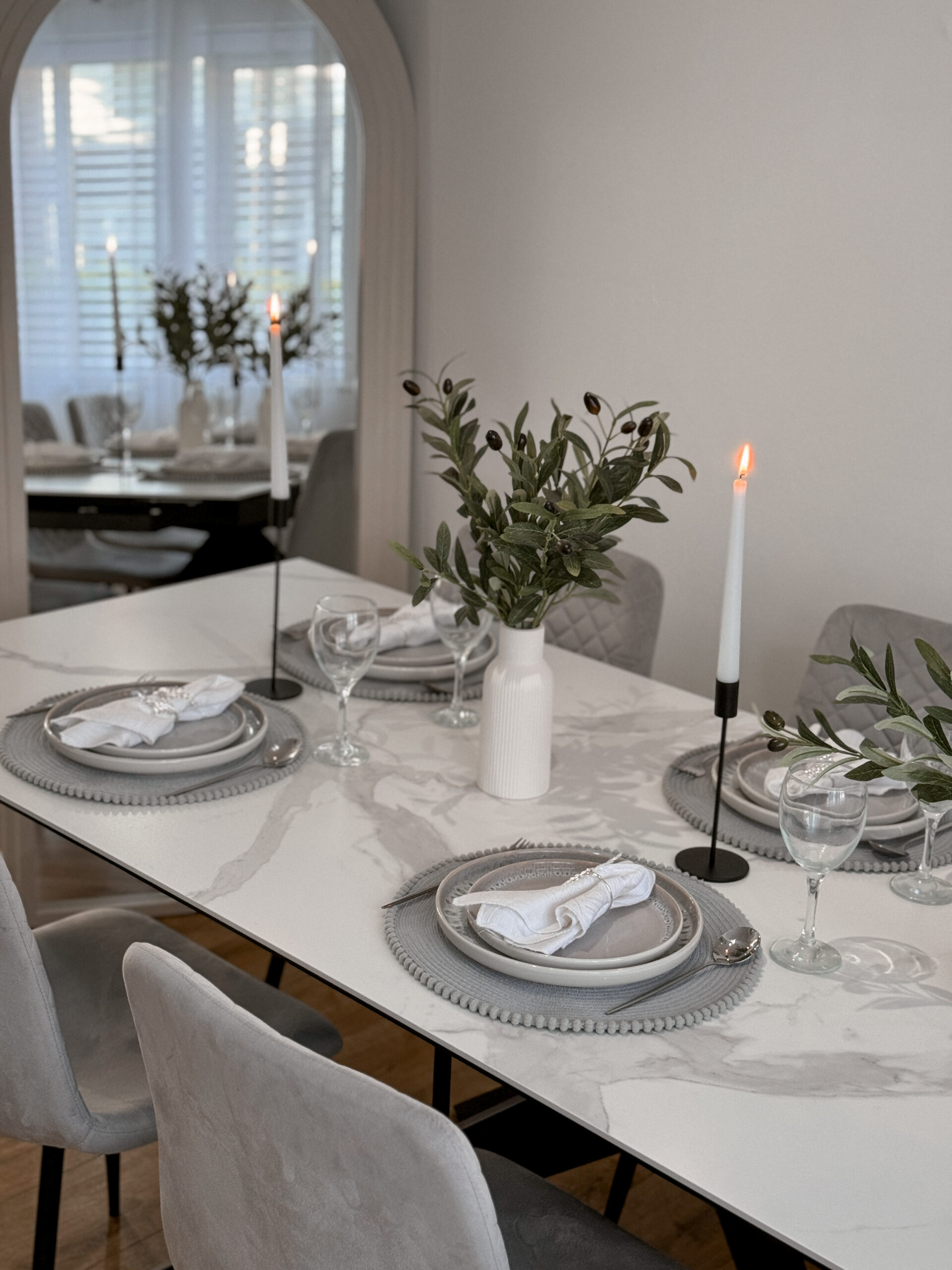 A dining table and chairs-dining room furniture-ceramic table-grey upholstered dining chairs-tapered candles-black candle holders-vases with foliage-arched mirror-grey place settings
