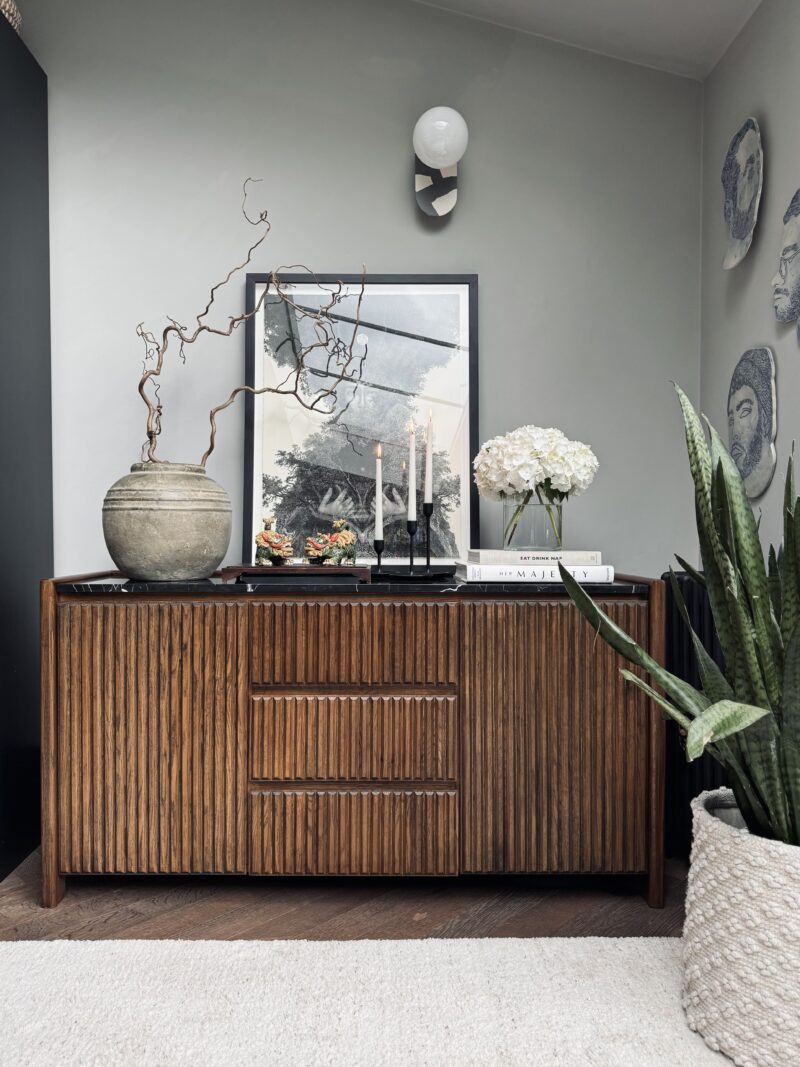 A sideboard-living room furniture-pale grey walls- dark wooden fluted sideboard with black marble top-black framed artwork-fresh white flowers-stacked books-tapered candles-large antique vase-indoor potted plant