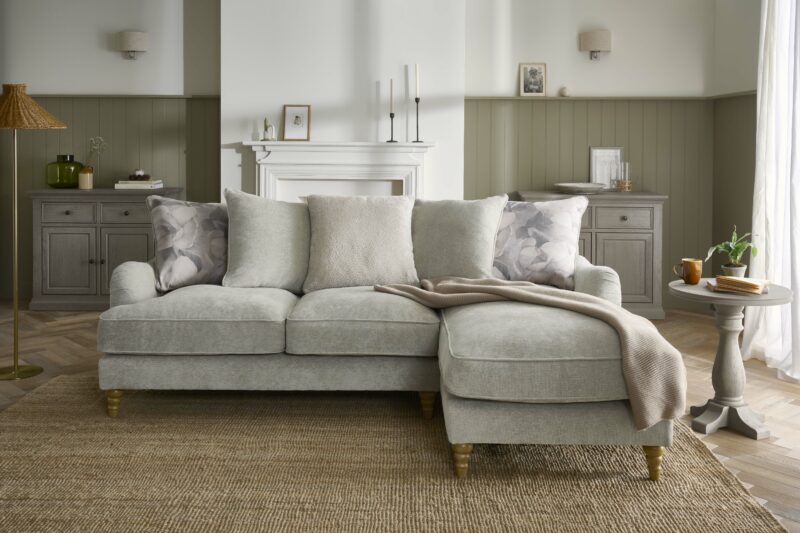 A sofa-living room furniture-grey corner chaise sofa-subtle floral cushions-jute rug-khaki green panelling-cream fireplace-blush through-grey wooden sideboards-rattan floor lamp-grey pedestal side table
