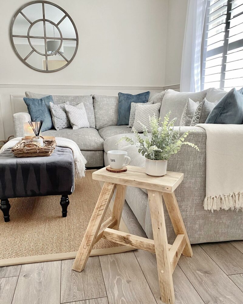 A modular corner sofa-living room furniture-pale grey corner sofa-denim blue accent cushions-dark grey footstool-rustic pale wooden stool-jute rug-cream walls-wooden floor-natural light