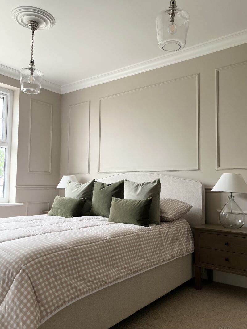 A bed and bedside table-bedroom furniture-neutral walls with beading detail-upholstered bed-layered gingham bedding-dark wood bedside table-brown carpet-white ceiling-statement vintage glass ceiling lights-glass bedside table lamp-natural light-ceiling rose