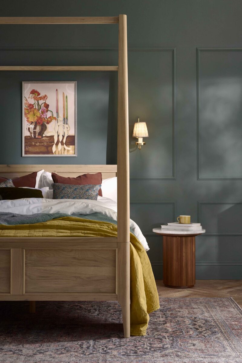 A bed and bedside table-bedroom furniture-four poster bed-fluted wood and marble side table-vintage style rug-parquet floor-round side table-moody blue walls-draped bed throw-layered bed linen- wall panelling-oversized artwork