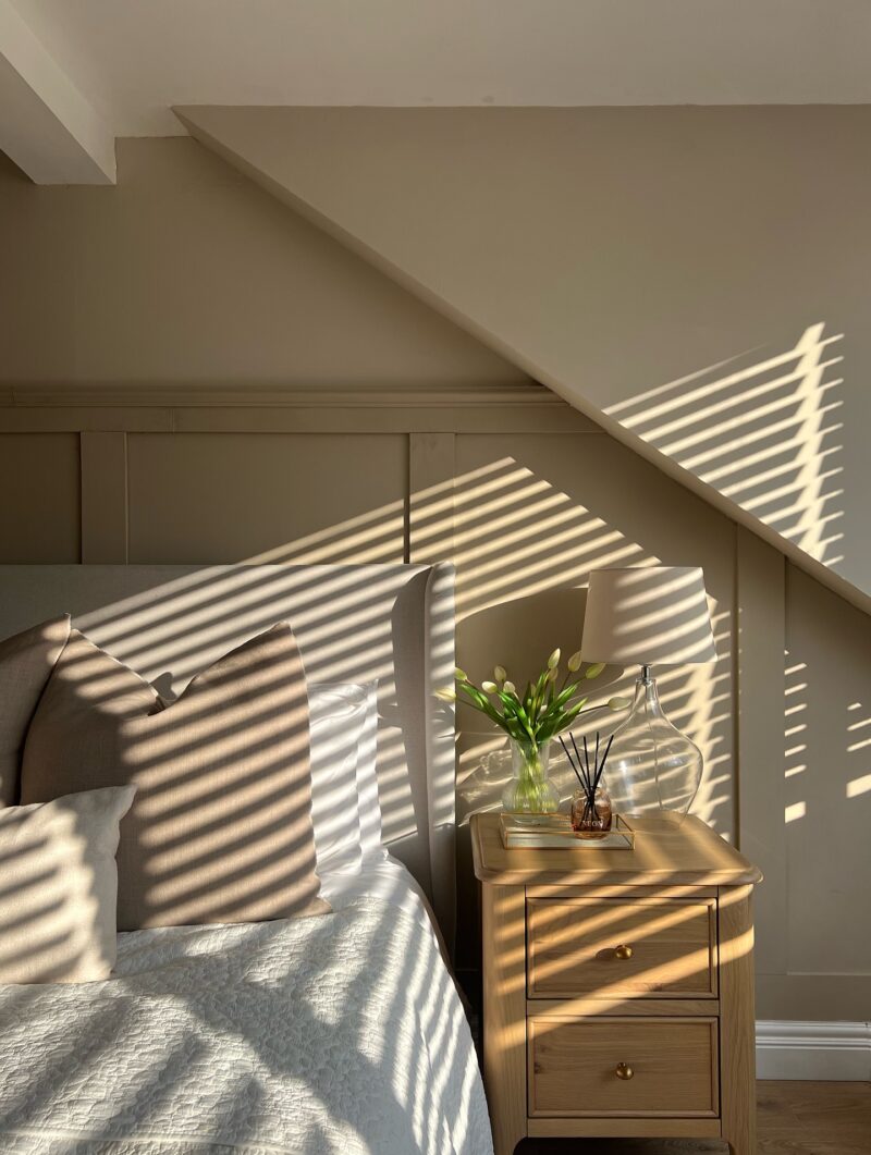 A bed and bedside table-bedroom furniture-beige panelled wall-upholstered bed-cosy neutral bedding-natural oak bedside table with drawers-fresh flowers in a glass vase-glass table lamp-scent diffuser-natural sunlight- shutter shadow