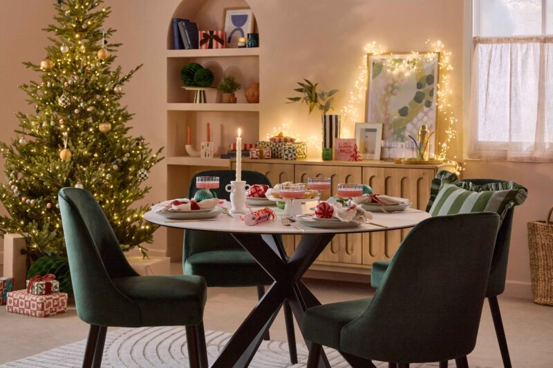 A dining table and chairs-dining furniture-white ceramic tabletop-green velvet dining chairs-stripey green cushion with frills-christmas tablescape-festive place settings-colourful baubles-striped crackers-twisted candles-scalloped champagne glasses-blush pink walls-fairy lights-christmas tree-oak wooden sideboard-arch shelving