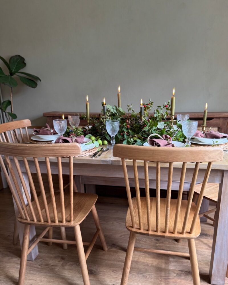 A dining table and chairs-dining room furniture-spindle back chairs-wooden table-autumnal tablescape-green tapered candles-table foliage-glassware-place settings-wooden floor-neutral walls