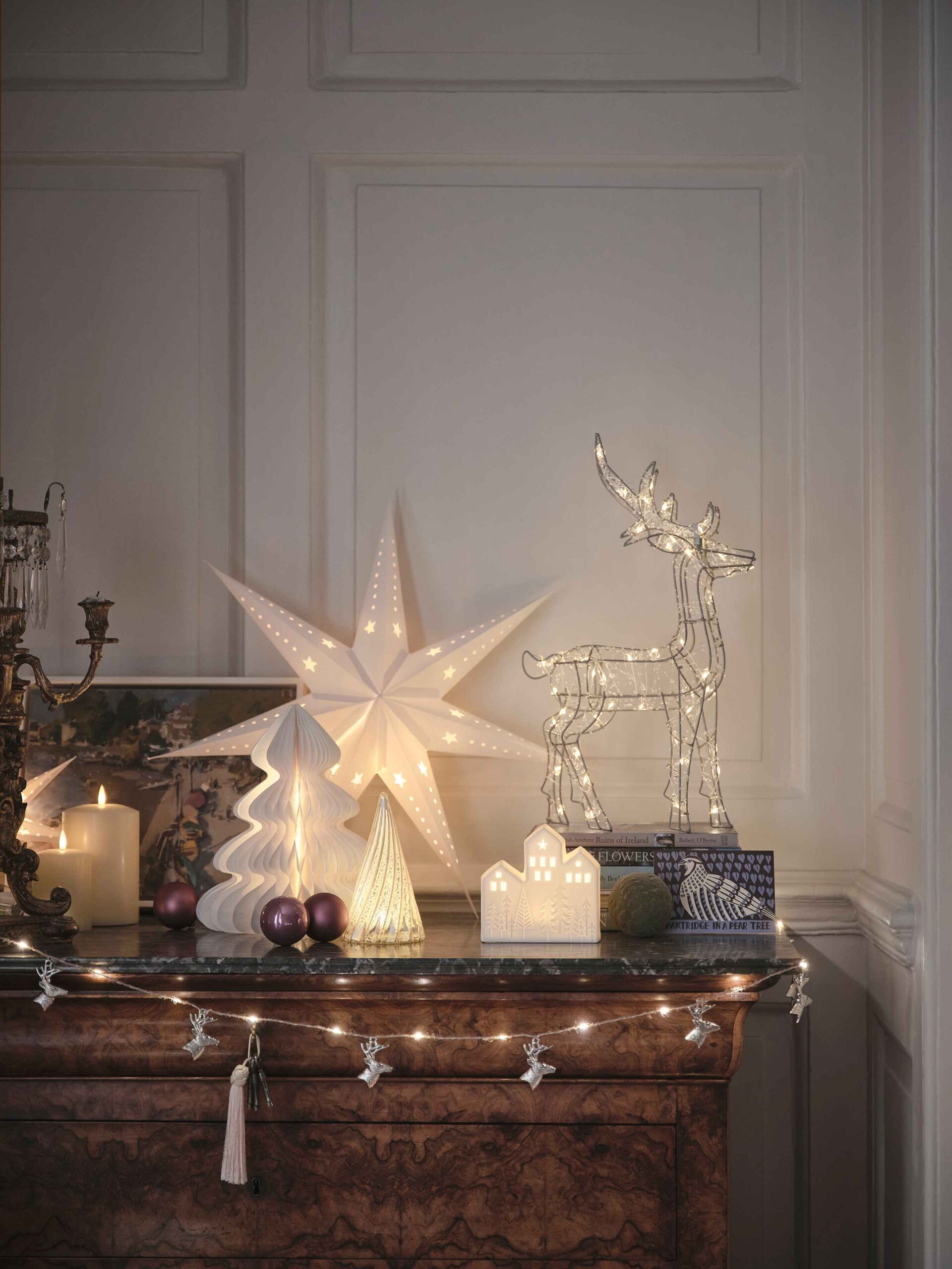 A sideboard-living room furniture-dark wooden sideboard-fairy lights-white star light-wire reindeer light-paper Christmas tree decorations-cream pillar candles-candelabra-painting-purple baubles-white panelled walls