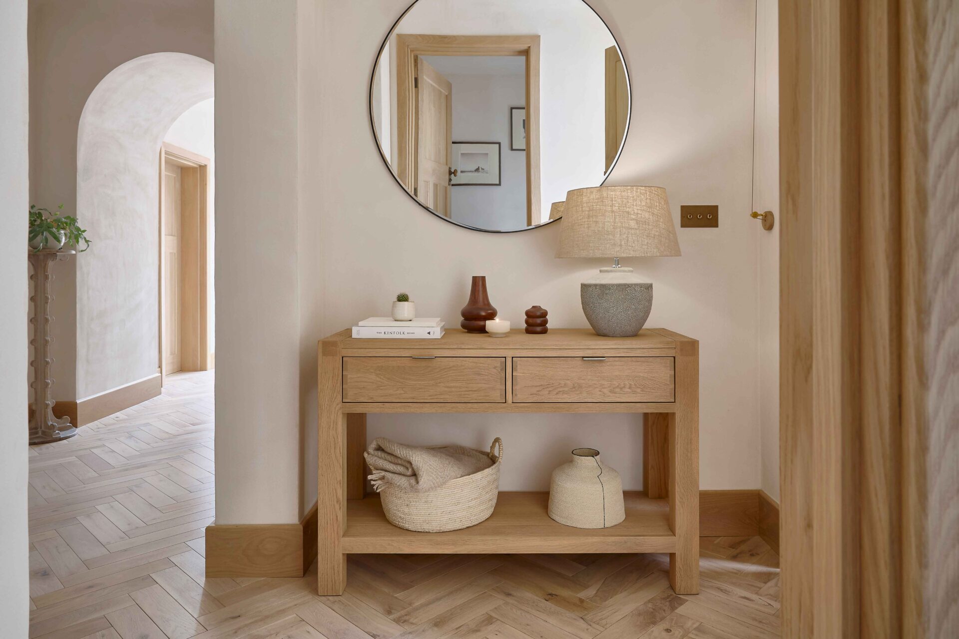 A console table-hallway furniture-parquet flooring-round mirror-textured table lamp-woven storage basket-sculptural vases-stacked books-cream walls-archway-entranceway