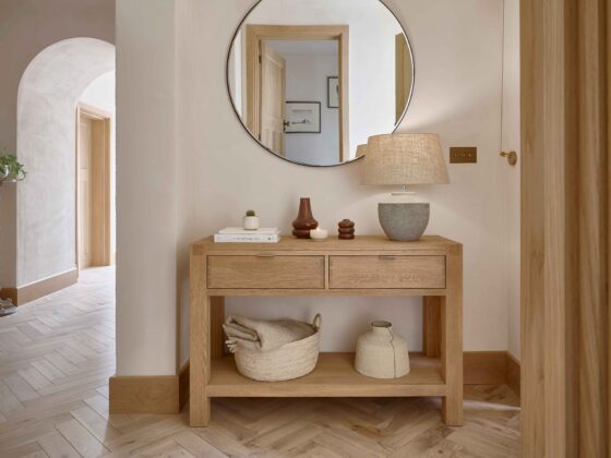 A console table-hallway furniture-parquet flooring-round mirror-textured table lamp-woven storage basket-sculptural vases-stacked books-cream walls-archway-entranceway