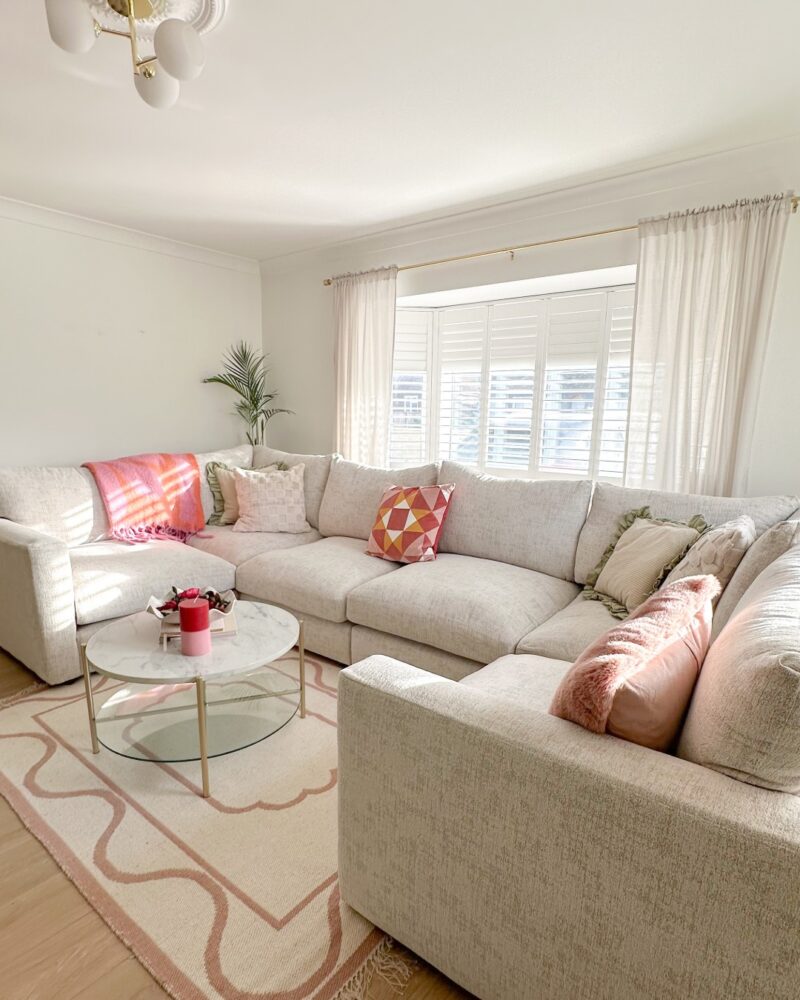 A corner sofa-living room furniture-off white walls-beige u-shaped sofa-cloud sofa-pink cushions-pink throw-neutral cushions-round marble coffee table-pink and cream rug-pale wooden floor-ceiling rose