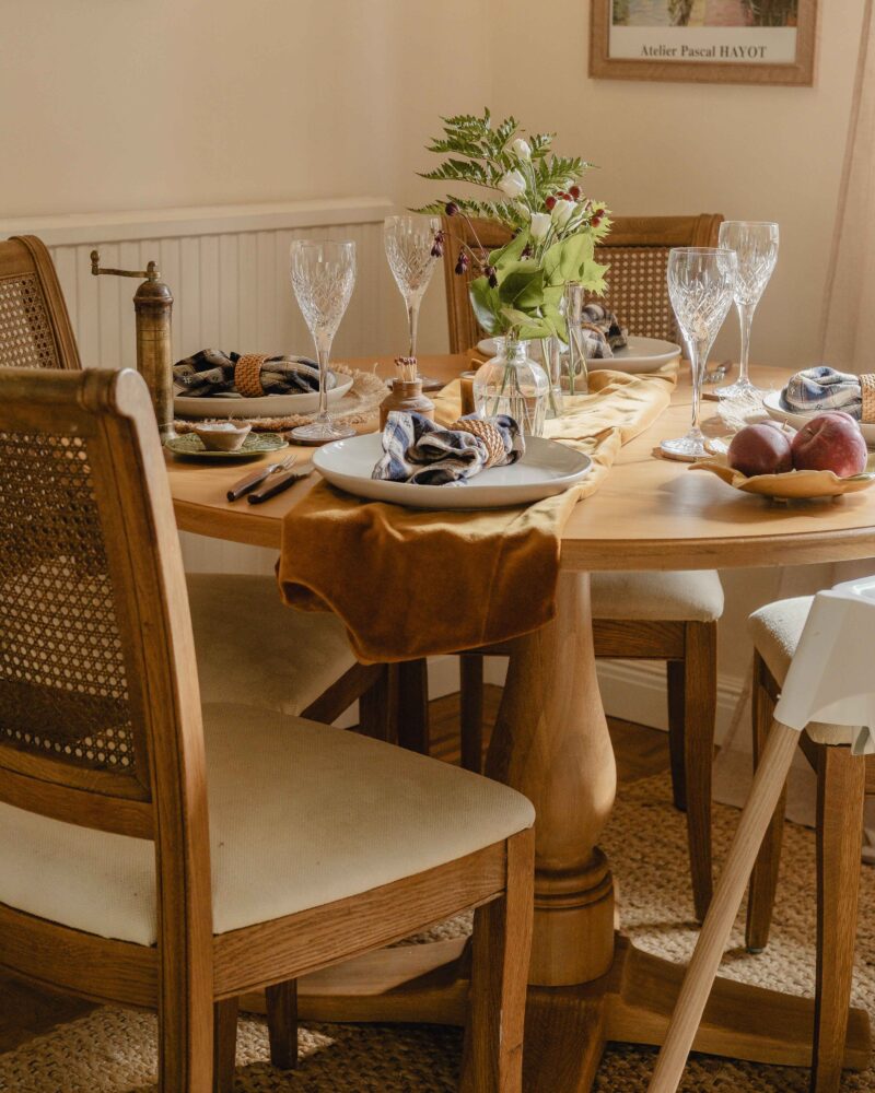 A dining table and chairs-dining room furniture-round wooden dining table-wooden upholstered dining chairs-pedestal base table-highchair-champagne glasses-white crockery-caramel fabric table runner-napkins and napkin rings-fresh foliage-neutral walls-jute rug-wooden floor-radiator