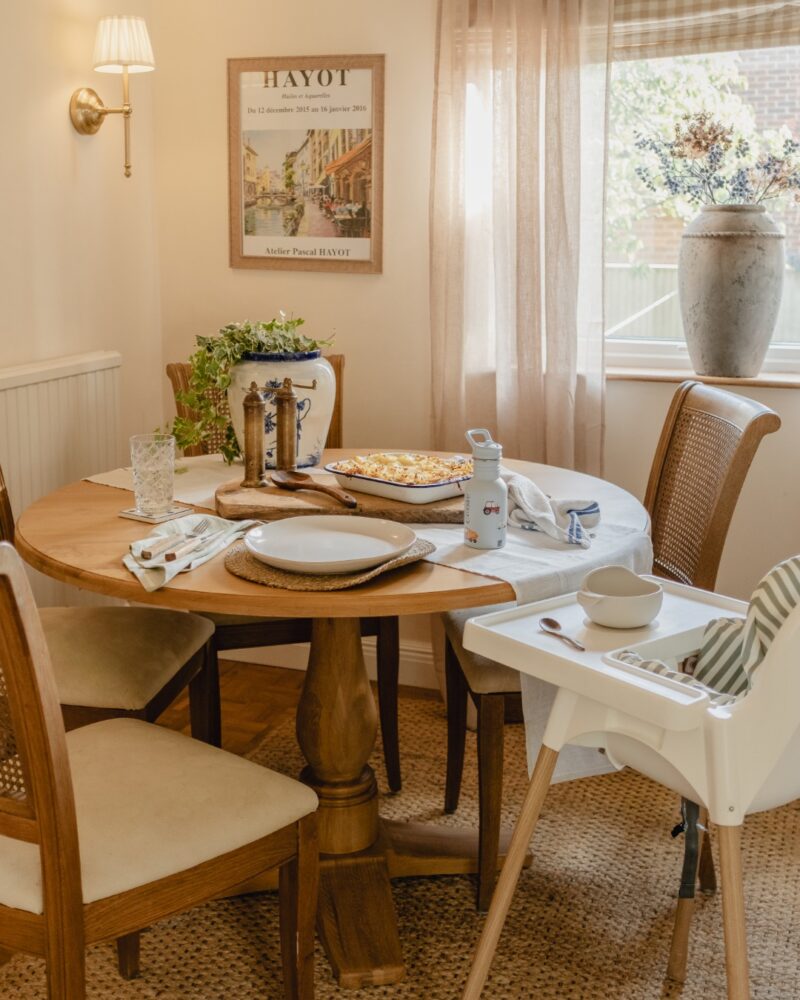 A dining table and chairs-dining room furniture-round wooden dining table-white high chair-wooden and fabric dining chairs-family table-wall art-wall lights-sheer curtains-neutral walls-house plant-vase of flowers