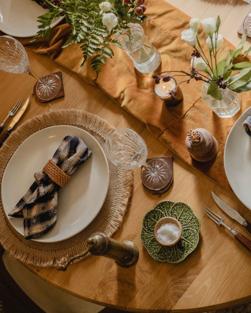 A table-dining room furniture-round wooden dining table-birds eye view-woven natural placemats-off white crockery-fabric table runner-fresh flowers in glass vases-crystal champagne glasses-matches pot-salt bowl-cabbage plate-pepper grinder-candle