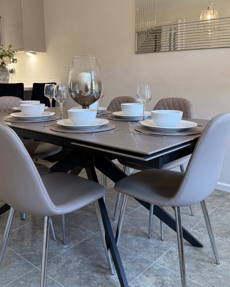 A dining table and chairs-dining room furniture-grey ceramic dining table-grey dining chairs-white crockery-wine glasses-neutral walls- large etched wall mirror-grey floor tiles-kitchen units