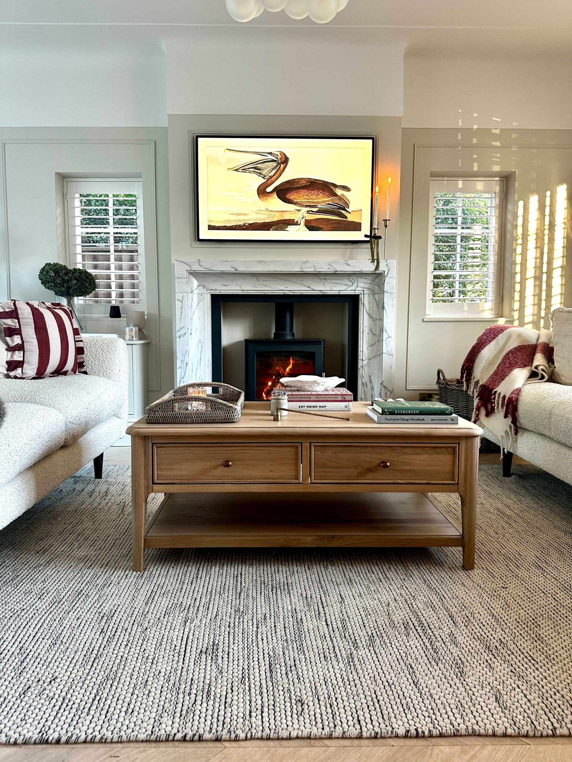 A coffee table and sofas-wooden coffee table with storage-textured neutral rug-marble fireplace-black log burner-cream sofas-striped red and cream cushion and throw-neutral walls-stacked books-rattan tray-art TV-window shutters