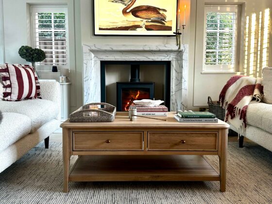 A coffee table and sofas-wooden coffee table with storage-textured neutral rug-marble fireplace-black log burner-cream sofas-striped red and cream cushion and throw-neutral walls-stacked books-rattan tray-art TV-window shutters