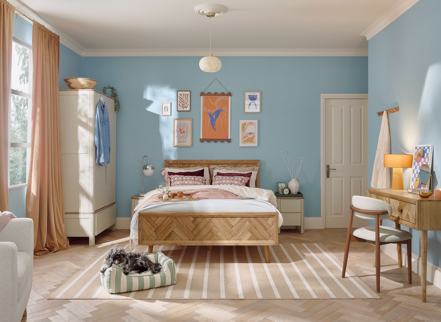 A bed, wardrobe, and dressing table-bedroom furniture-wooden double bed-neutral painted double wardrobe-sky blue walls-striped natural rug-wooden dressing table-dark wood upholstered mid century style chair-white bedroom door-art wall-striped dog bed-grey dog-parquet wooden floor-draped curtains-cream armchair