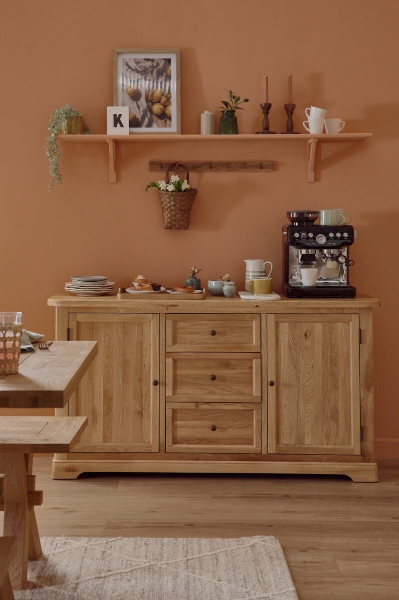 A sideboard-dining room furniture-natural oak dining table and benches-soft terracotta walls-neutral rug-large wooden sideboard-black coffee machine-shelving-peg shelf-woven hanging basket-stacked plates-pastries-tapered candles-framed print-crockery-house plant