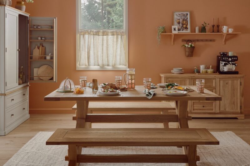 A dining table set-dining room furniture-natural oak dining table and benches-soft terracotta walls-neutral rug-large wooden sideboard-neutral painted wooden larder-cafe curtain-spring summer tablescape-coffee machine-shelving