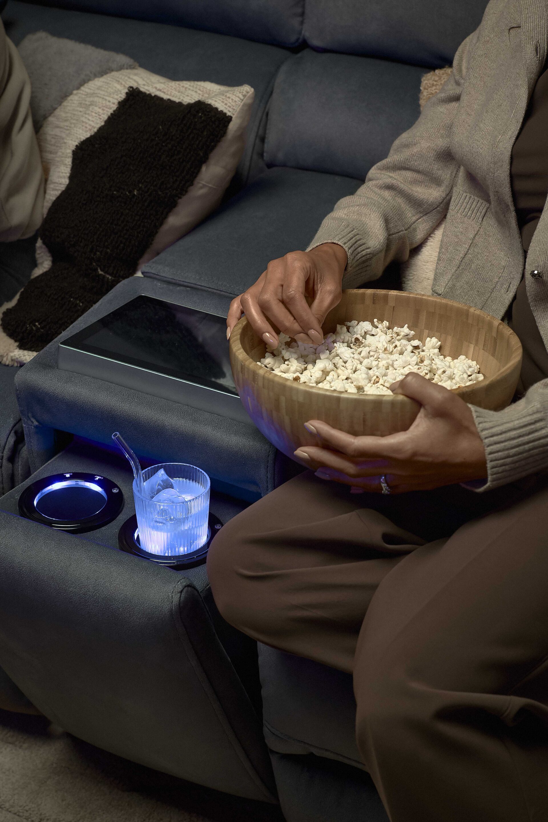 A sofa-living room furniture-dark blue recliner sofa with console screen and cup holders-tactile cushion-armrest-iced drink in glass cup with straw-person eating popcorn-wooden bowl-neutral clothing