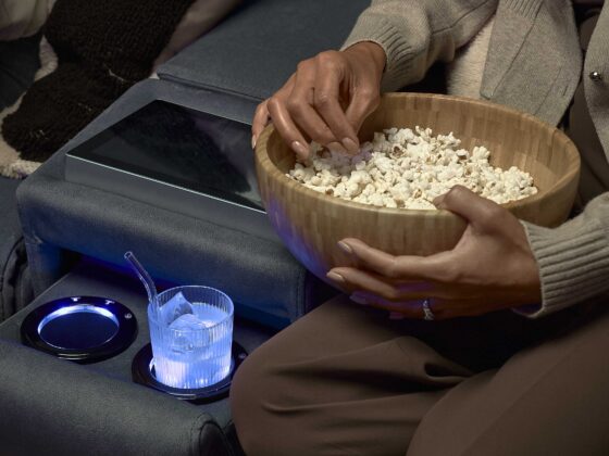 A sofa-living room furniture-dark blue recliner sofa with console screen and cup holders-tactile cushion-armrest-iced drink in glass cup with straw-person eating popcorn-wooden bowl-neutral clothing